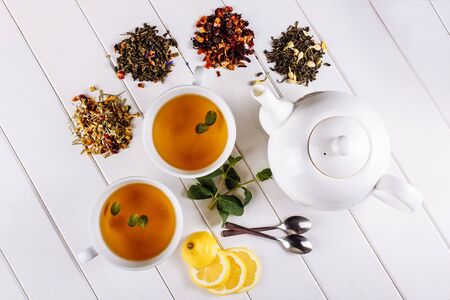 Different sorts of dry tea on white wooden background and two cups and pot. Top view decorated with mint and lemonの写真素材