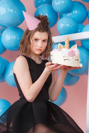 Happy teen girl holding unicorn layered cake decorated with meringues close-up. Pink background with blue balls.の写真素材