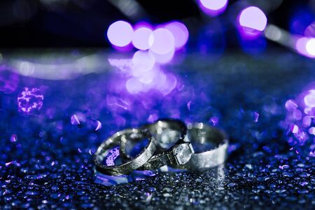 wedding and engagement rings of white gold or silver on sparkling purple wet background with water drops. Copy space for invitation card text.の写真素材