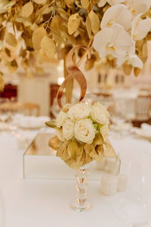 Decorative Bouquet Souvenir on Crystal Stem Photo. Elegance Floral Ornament Made from White Rose Buds, Golden Leaves Standing on Dinner Table. Luxury and Aromatic Decor. Blurred Backgroundの写真素材