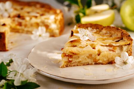 Apple Pie Sliced Piece with Crispy Crust Snack Close-up Photo. Homemade Fresh Bakery Cake Food Ornamented Blossom Flowers, Fruit and Green Leaves Branch on Blurred Background. Tasty Nutrient Breakfastの写真素材