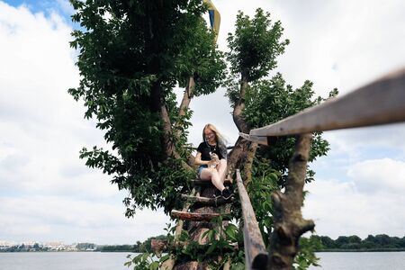 A young woman with a three-month-old Scottish Straight kitten sits on a beautiful green tree above the lake in summer. Walk, rest with a pet. The blonde is wearing short denim shorts, a black cotton T-shirt and glasses.の写真素材