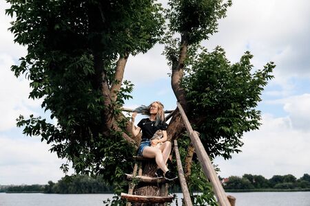 A young woman with a three-month-old Scottish Straight kitten sits on a beautiful green tree above the lake in summer. Walk, rest with a pet. The blonde is wearing short denim shorts, a black cotton T-shirt and glasses.の写真素材