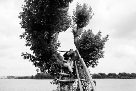 A young woman with a three-month-old Scottish Straight kitten sits on a beautiful green tree above the lake in summer. Walk, rest with a pet. The blonde is wearing short denim shorts, a black cotton T-shirt and glasses.の写真素材