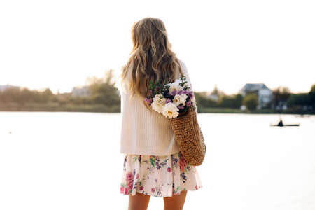 This stock photo shows a back young blonde woman walking on the beach in a dress and sweater with straw bag and flowers on a sunset.の写真素材