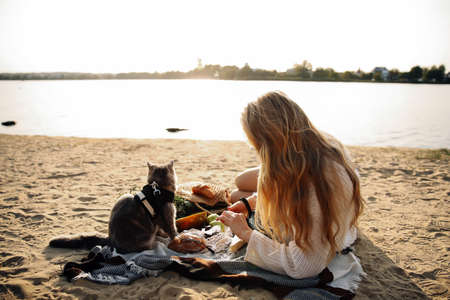 Young blonde woman lying on the beach in autumn in a white knitted sweater with a cat on a sunny day...