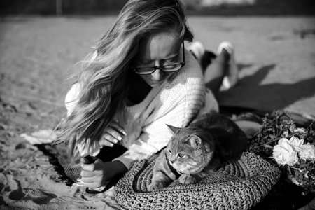 Top view of a woman in glasses and a white broadcast sweater lying on the beach with her kitten breed Scottish Rectangular gray at sunset. The cat lies on a straw bag near the flowers.の写真素材