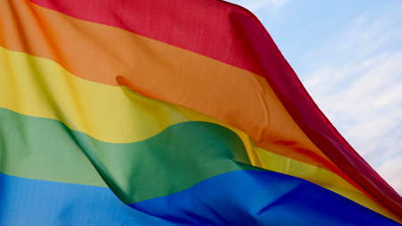 Female hand holding a LGBT gender identity flag on a background of blue cloudy sky on a sunny windy summer day during the celebrating a gay parade, Bisexuality or Coming Out Dayの写真素材