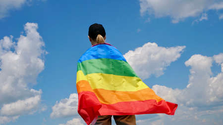 Bisexual, lesbian, woman, transgender stands with LGBT flag against blue sky with clouds on a sunny...