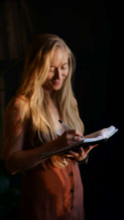 Blond caucasian woman in dress writes in a notebook on a dark black background on a sunny day. The young secretary is working. Vertical photo in defocusの写真素材