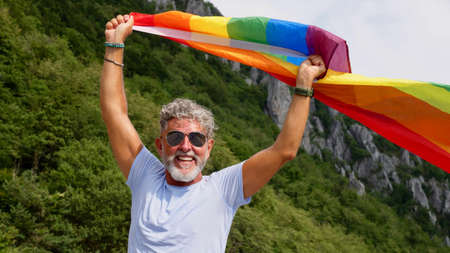 Portrait of a gray-haired senior elderly Caucasian man bisexuality with a beard and sunglasses holding a rainbow LGBTQIA flag on nature. Celebrates Pride Month, Rainbow Flag Day, gay paradeの写真素材