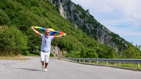 Portrait of a gray-haired senior elderly Caucasian man bisexuality with a beard and sunglasses holding a rainbow LGBTQIA flag on nature. Celebrates Pride Month, Rainbow Flag Day, gay paradeの写真素材