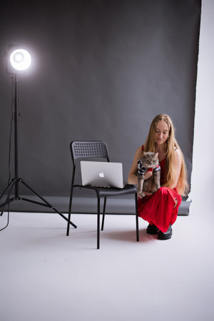 A business woman, freelancer, blogger in a red dress is working, typing on a laptop apple on a white floor, black background. blonde assistant in video production with computer MacBook and catのeditorial素材