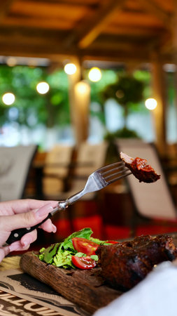 A persons hand holds a fork with a piece of roasted meat, ready to eat. The warm restaurant atmosphere is visible in the blurry background, highlighting a delicious meal.の写真素材
