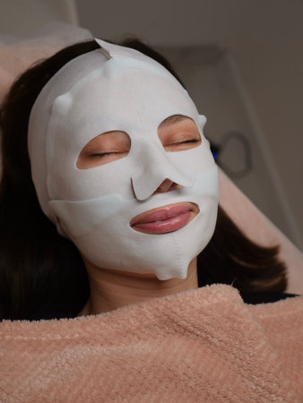 A relaxed young woman lies on a spa bed receiving a sheet mask treatment for hydration and rejuvenation. Her eyes are closed in peaceful tranquility under professional careの写真素材