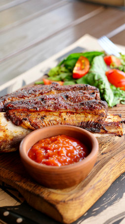 A vertical close-up of delicious grilled ribs served on a rustic wooden board. The dish is accompanied by a bowl of rich sauce and a fresh salad, creating a mouthwatering meal.の写真素材