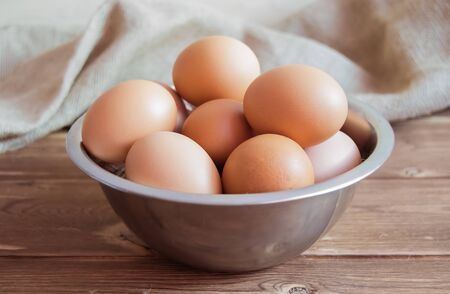 Chicken eggs in a metal bowl on a wooden background with a napkin