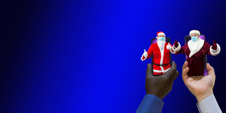 Afroamerican and caucasian-white hands holding phones with 2 Santa Clauses. Santa Clauses have medical masks on. Close-up on a blue gradient background.の写真素材
