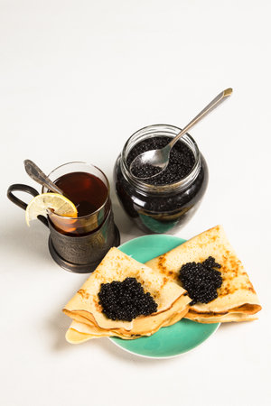 Traditional Russian pancakes with black caviar. In the background is a jar with sturgeon caviar and a glass in a cup holder with black tea.の写真素材