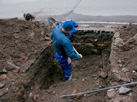 A man handyman digs a trench in the winter in the city by hand with a shovel. Construction preparatory work.の写真素材