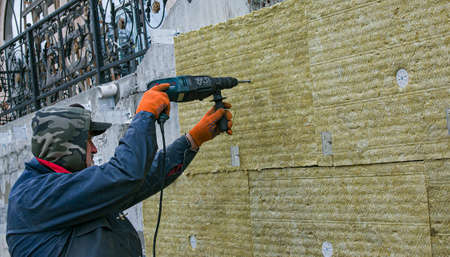 Dnepropetrovsk, Ukraine - 04/06/2021: A man installs a layer of thermal insulation on the wall of the stairs from the outside - using mineral wool panels. Working with a drill. The concept of repair and insulation of external structures.のeditorial素材