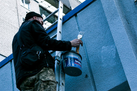 Dnepropetrovsk, Ukraine - 03.29.2021: The work of the painter is on top. An employee paints the facade of a building while standing on the stairs.のeditorial素材