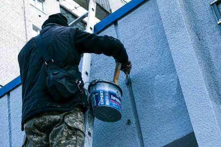 Dnepropetrovsk, Ukraine - 03.29.2021: The work of the painter is on top. An employee paints the facade of a building while standing on the stairs.のeditorial素材