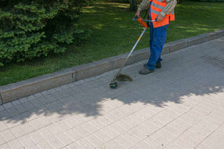 A worker in overalls with a trimer in his hands mows the grass on the lawn and paths in the park....