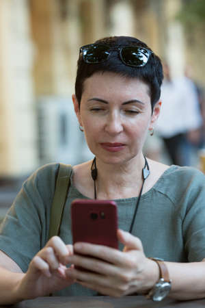Attractive middle-aged woman with a phone in the city. A woman sits at a table and looks through...