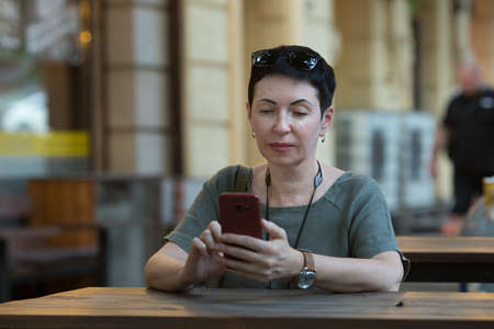 Attractive middle-aged woman with a phone in the city. A woman sits at a table and looks through...