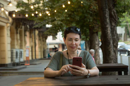 Attractive middle-aged woman with a phone in the city. A woman sits at a table and looks through messages on the phone.の写真素材