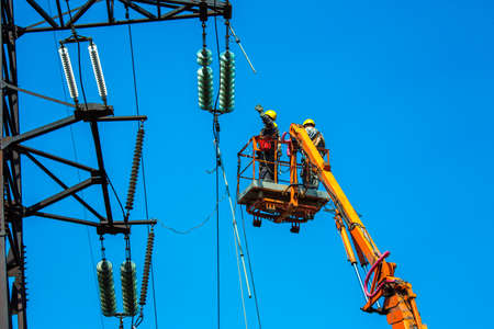 High voltage power line transmission tower workers with crane and blue sky. Hydro linemen on boom lifts working on high voltage power line towers.の写真素材