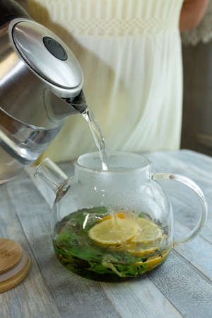 A woman pours hot water into a kettle with mint, tea and sliced lemon. The process of making tea...