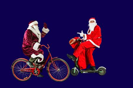 Two merry Santa Clauses in Christmas hats and red suits ride a scooter and a bicycle with bags of...