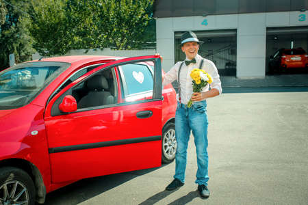 A young man in a hat gets out of the car. In the hands of a man is a bouquet of white-yellow flowers.の写真素材