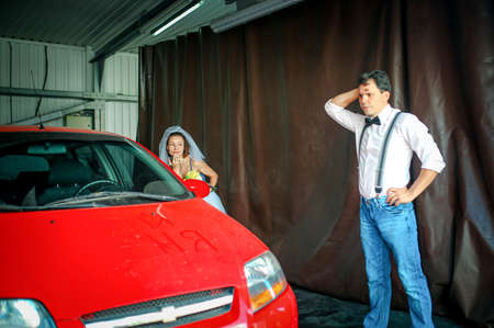 A young man with a bow tie and a woman in a veil are washing a car at a car wash. wedding anniversary. Humor.の写真素材