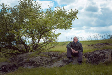 Portrait of a middle-aged man tourist sitting on a large stone on the bank of a narrow river....