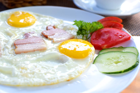 Fried eggs with pieces of bacon, tomatoes and cucumbers. Breakfast on a white plateの写真素材