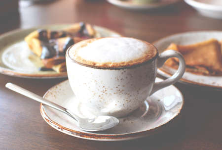 Coffee in a beautiful ceramic cup on a wooden table in a cafe on the background of plates with pancakes. Cappuccino coffee.の写真素材