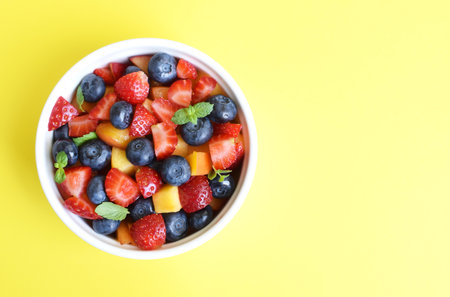 Fruit salad in a white cup on a yellow background. Sliced strawberries, peaches and blueberries with mint leaves. Free space for text.の写真素材