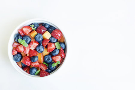 Fruit salad in a white cup on a white background. Sliced strawberries, peaches and blueberries with mint leaves. Free space for text.の写真素材