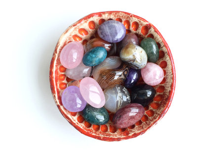 Colorful tumbled semi-precious stones lie in a handmade ceramic bowl. Red bowl with agate, rose quartz, amethyst and apatite on a white background.の写真素材