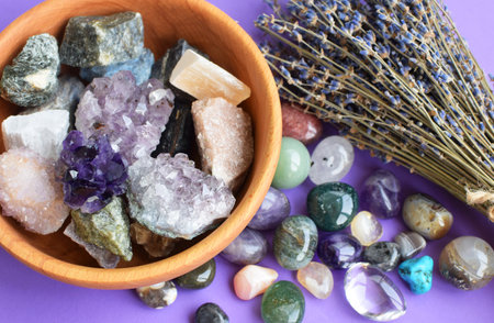 Semi-precious stones in a wooden bowl with a dry lavender bouquet. Amethyst and amethyst druse crystals, rose quartz, agate, apatite, aventurine, turquoise, rock crystal on a purple background.の写真素材