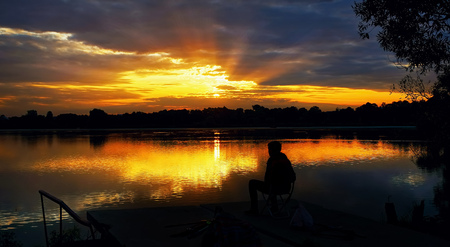 The silhouette of a seated fisherman on a beautiful background of a rising sun with rays in the dramatic clouds .の写真素材