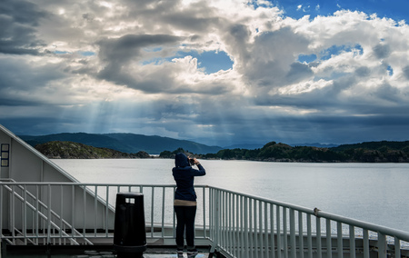 Silhouette of woman in hooded jacket on the deck of the ferry taking pictures of the beautiful Northern landscape with sun rays passing through cloud in the North sea..の写真素材