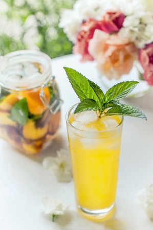 Transparent glass with water, detoxication with pieces of peach and apricot, with ice and mint close- up. A refreshing drink on the background of bouquet of pink peonies.の写真素材
