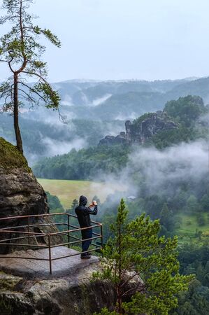 The silhouette of a person with a phone is standing with his back to the photographer on the rock and looking at the mountains , sky and clouds on a rainy summer day. The vertical frame.の写真素材