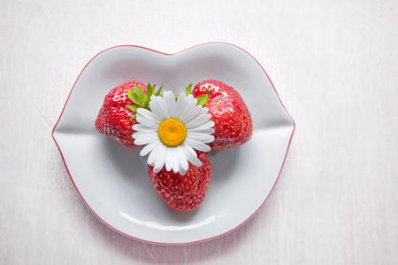 Ripe strawberry and a Daisy flower are on a white plate in the shape of lips on a light wooden background. Copy space. Festive greeting. The horizontal frame.の写真素材