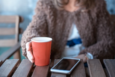 Hands using a phone texting on smartphone app and holding paper cup of coffee.