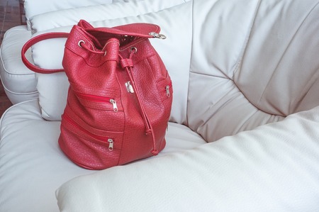 A woman's coral-colored backpack stands on a leather white sofa in the hall. Things to travel.の写真素材